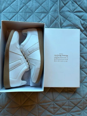 Maison Margiela Gats - Säljer ett par stilrena vita sneakers från Maison Margiela med beige detaljer och gummisula. Skorna har en mix av slätt läder och mocka, klassisk låg profil och vita skosnören. Perfekta för dig som gillar minimalistisk och tidlös design. Helt oanvända
