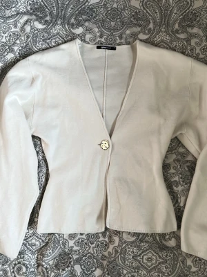 Vit kofta från Gina Tricot - Jättefin vit kofta från Gina Tricot med djup V-ringning. Koftan går inåt i midjan och är lite bredare vid axlarna. Använd endast få gånger.