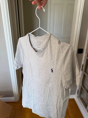 Ljusgrå t-shirt från Ralph Lauren - Snygg ljusgrå t-shirt från Ralph Lauren med klassisk blå logga broderad på bröstet. T-shirten har rund hals och korta ärmar, perfekt för en clean och enkel stil. Tillverkad i mjuk bomull som känns skön mot huden.