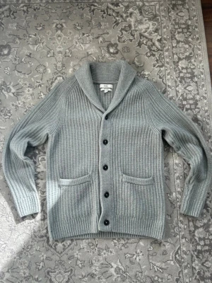 Grå ribbstickad kofta  - Snygg grå ribbstickad kofta med sjalkrage och fem svarta knappar framtill. Koftan har två fickor fram och är gjord i ett mjukt, tjockt material som känns riktigt lyxigt. Perfekt för lager på lager och chill stil.