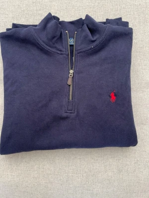 Marinblå half-zip tröja från Polo Ralph Lauren - Klassisk marinblå tröja från Polo Ralph Lauren med half-zip och hög krage. Tröjan har det ikoniska röda Polo-logotypen broderat på bröstet och är tillverkad i mjukt bomullsmaterial. Perfekt för en stilren och avslappnad look.