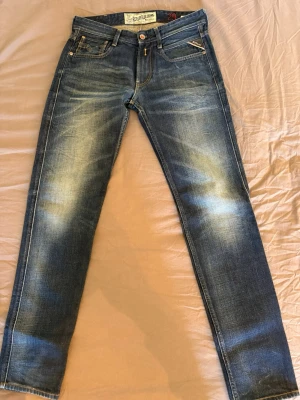 Blå slim fit jeans från Replay W29 L32 - Säljer ett par blå slim fit jeans från Replay med snygga slitningar och klassisk femficksdesign. Jeansen har en något tvättad look och smal passform som sitter tajt längs benen. Perfekta för dig som gillar en modern och avslappnad stil.