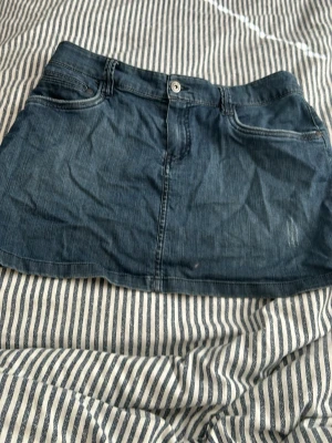 Mörkblå jeanskjol med fickor - Snygg mörkblå jeanskjol i kort modell med klassiska fickor både fram och bak. Kjolen har knapp och dragkedja framtill och är gjord i ett slitstarkt denimtyg. Perfekt att matcha med en enkel t-shirt eller hoodie.