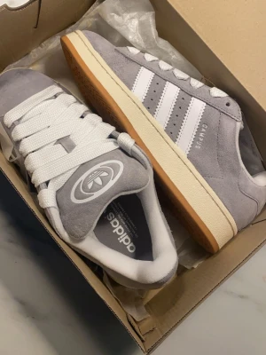 Adidas Campus grå sneakers - Snygga Adidas Campus sneakers i grå mocka med vita detaljer och klassiska tre ränder på sidan. Skorna har vit sula med gummiyta undertill och breda vita skosnören. Perfekta för en avslappnad streetwear-look.