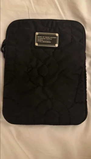 Svart ipadfodral Marc by Marc Jacobs - Snygg svart ipadfodral från Marc by Marc Jacobs med quiltat mönster och metallbricka framtill. Väskan har en mjuk, vadderad känsla och dragkedja längs kanten. Perfekt för att bära din laptop med stil.