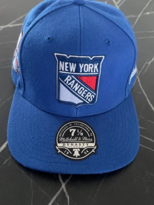 Blå New York Rangers keps - Snygg blå keps från Mitchell & Ness med New York Rangers-logga framtill och ett broderat 75-årsjubileumsmärke på sidan. Klassisk rak skärm och broderade ventilationshål. Perfekt för dig som gillar hockey och sportig stil.