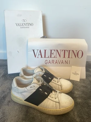 Valentino Skor - Snygga vita sneakers från Valentino med bred svart läderdetalj över mitten och ikoniska nitar på hälen. Skorna har klassisk låg profil, vita snören och diskret logga på plösen. Tillverkade i slätt läder för en clean och modern look.