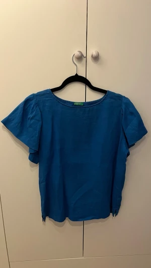Blå kortärmad blus - Snygg blå blus från United Colors of Benetton med rund halsringning och korta, löst sittande ärmar. Tillverkad i linne ett lätt och luftigt tyg som ger en avslappnad känsla. Perfekt att matcha med jeans eller kjol för en trendig look.