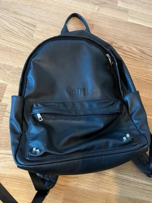 Svart ryggsäck från YOURTURN - Snygg svart ryggsäck i fejkläder med silverfärgade detaljer framtill. 