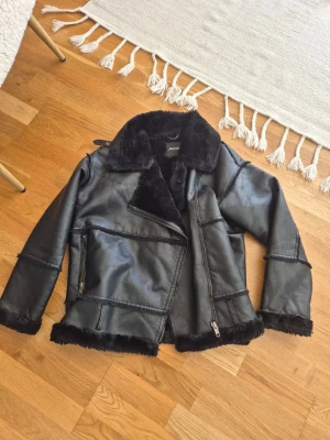 Svart skinn pälsjacka från Monki - Säljer en svart pälsjacka från Monki med bred krage och dragkedja framtill. Jackan har en oversized passform, synliga sömmar och mjukt foder i fuskpäls. Perfekt för kalla vinterdagar och ger en cool look till din outfit.