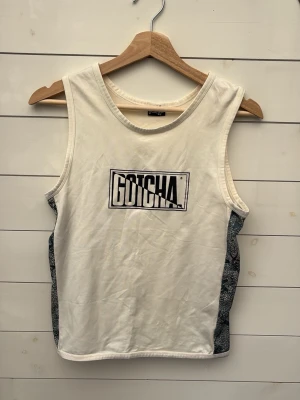 Y2K Vit linnetopp från Gotcha - Säljer en vit linnetopp från Gotcha med tryckt logga framtill. Toppen är ärmlös och har kontrasterande sidopaneler i grått mönster. Perfekt för dig som gillar en sportig och avslappnad stil.