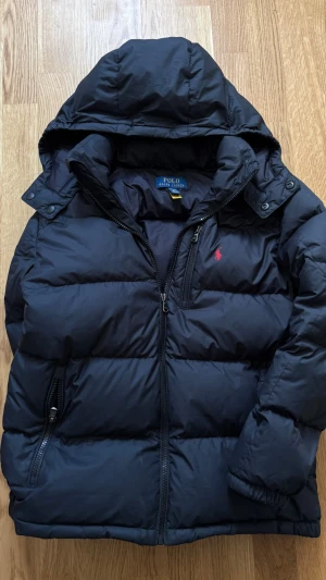 Svart dunjacka från Polo Ralph Lauren - Snygg svart dunjacka från Polo Ralph Lauren med avtagbar huva och dragkedja framtill. Jackan har två sidofickor med dragkedja och den klassiska röda loggan broderad på bröstet. Perfekt för kalla vinterdagar och riktigt stilren. OBS! Fickorna sitter fast (går att fixa väldigt lätt)