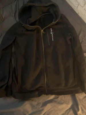 Svart hoodie från Sail Racing - Svart hoodie från Sail Racing med dragkedja framtill och en bröstficka med dragkedja. Märkets logga är tryckt på bröstet. Hoodien har en klassisk huva och ribbade muddar. Perfekt för en chill och sportig stil.