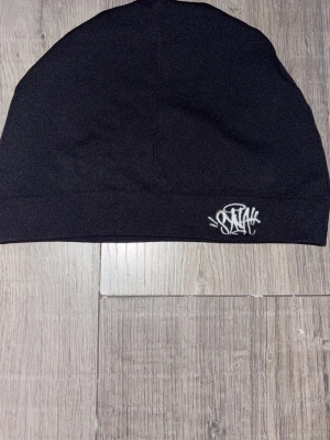 Svart syna world skull cap med vit logo - Säljer en svart syna world skull cap med en vit broderad logga framtill i streetstil. Mössan är enkel och stilren, tillverkad i mjukt stretchigt material som sitter skönt på huvudet. Perfekt accessoar för dig som gillar en clean look.