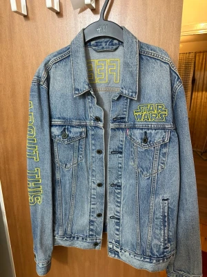 Star Wars jeansjacka från Levi's - Blå jeansjacka från Levi's med Star Wars-tryck i gult på bröstet och texten 'BAD FEELING ABOUT THIS' på ryggen och ärmen. Klassisk modell med bröstfickor och knappar framtill. Perfekt för dig som vill sticka ut med en unik och nördig stil.
