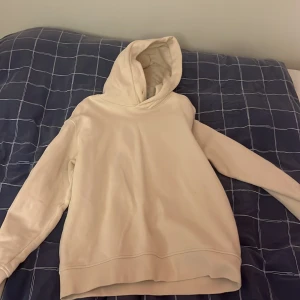 Beige basic hoodie utan tryck - En stilren beige hoodie med huva och ribbade muddar. Hoodien är helt utan tryck eller logga och har en avslappnad passform. Perfekt för dig som gillar en clean och enkel look. Tillverkad i mjukt sweatshirtmaterial.