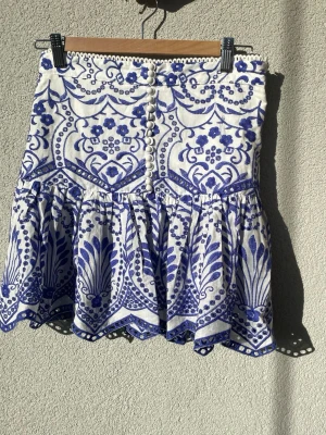 Slutsåld kjol från Zara - Supersöt kortkjol i vitt och blått med volangkant och dekorativa knappar framtill. Kjolen har ett detaljerat blommigt mönster och är gjord i ett lätt bomullstyg, perfekt för varma dagar. Slutsåld på hemsidan och passar perfekt till sommaren 🍸