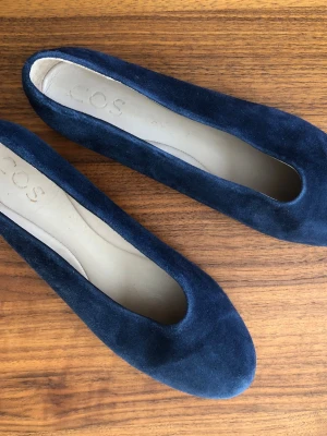 Mörkblå ballerinaskor från COS - Säljer ett par stilrena mörkblå ballerinaskor från COS i mjuk mocka. Skorna har rundad tå och en klassisk, enkel design som passar till det mesta. Insidan är ljus och skön för foten. Perfekta för dig som gillar minimalistisk stil.