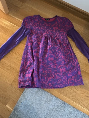 Lila långärmad tröja med blommor - Långärmad tunikaliknande tröja i lila med rosa blommigt mönster över hela plagget. Rund halsringning och rynk under bysten ger en avslappnad passform. Ärmarna är enfärgade medan kroppen har ett detaljerat tryck. Troligen i mjuk bomullsblandning. Är lite tajt. Kontakta för fler frågor 💕