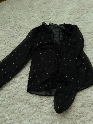 Svart prickig blus med långa ärmar - Snygg svart blus i transparent material med vita prickar och långa ärmar. Blusen har smock upptill, volang vid halsen och knytband framtill. Perfekt för dig som gillar trendiga detaljer och luftig känsla.