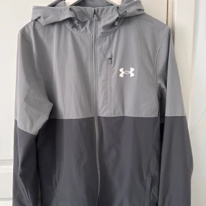 Grå vindjacka från Under Armour.  - Snygg tvåfärgad grå vindjacka från Under Armour med huva och dragkedja hela vägen. Jackan har en vit logga på bröstet, praktisk bröstficka med dragkedja och sportig look. Perfekt för blåsiga dagar och outdoor-aktiviteter. Pyttelitet svart märke där bak. Och litet hål i fickan 