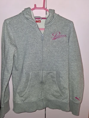 Grå zip hoodie från Puma - Snygg grå hoodie från Puma med dragkedja framtill och rosa logga på bröstet. Mjuk insida, två fickor fram och klassisk huva. Perfekt för chill dagar eller till träningen. Ribbstickade muddar vid ärmslut och nederkant. Materialet känns som bomullsblandning.