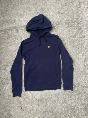 Mörkblå hoodie från Lyle & Scott - Snygg mörkblå hoodie från Lyle & Scott med klassisk gul logga på bröstet. Hoodien har huva med dragsko och en stor magficka framtill. Tillverkad i mjukt bomullsmaterial och har ribbade muddar vid ärmslut och nederkant.