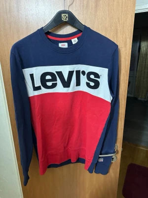 Blå och röd stickad tröja från Levi's - Snygg långärmad stickad tröja från Levi's i marinblått och rött med vit panel och stor svart logga framtill. Rund halsringning och ribbade muddar. Perfekt för dig som gillar streetwear och vill ha en klassisk look.