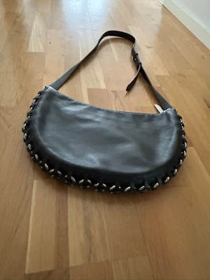 Michael Kors Svart axelväska med kedjedetaljer - Michael Kors. Cool svart axelväska i halvmåneformad design med justerbart axelband. Väskan är gjord i slätt läder och har snygga silverfärgade kedjedetaljer längs kanten som ger en edgy vibe. Perfekt för dig som vill sticka ut lite extra.