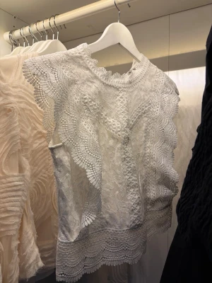 Vit somrig spetsblus  - Supersöt vit blus i spets med dekorativa volangkanter och rund hals. Blusen är ärmlös och har ett romantiskt mönster i spetsmaterial som ger en lyxig känsla. Perfekt att styla med jeans eller kjol för en trendig look.