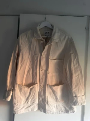 Vit overshirt från Zara - Stilren vit overshirt från Zara i kraftigt bomullstyg. Modellen har tre stora utanpåfickor framtill, knäppning med vita knappar och klassisk krage. Perfekt lager-på-lager-plagg med avslappnad passform och raka ärmar.