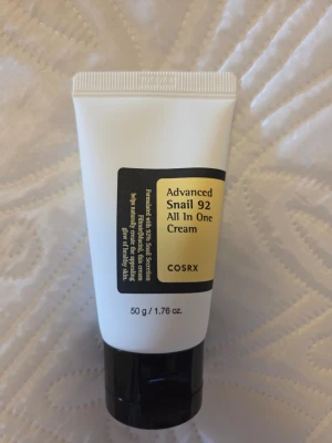 COSRX Advanced Snail 92 All In One Cream - En multifunktionell ansiktskräm från COSRX med snigelsekretfiltrat som återfuktar och stärker huden. Tuben är vit med svart och gul etikett och har svart lock. Innehåller 50 g och passar för dig som vill ha glow och mjuk hud. Endast testad en gång.
