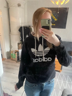 Svart Adidas hoodie med stor logga - Säljer en svart hoodie från Adidas med stor vit Trefoil-logga och text på bröstet. Hoodien har huva med vita dragsnören och en klassisk känguruficka framtill. Perfekt för en avslappnad och sportig stil.