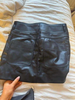 Svart kortkjol i skinnimitation - Snygg svart kortkjol i skinnimitation med stora fickor både fram och bak. Kjolen har silverfärgade knappar på framfickorna och dragkedja framtill. Perfekt för en trendig och edgy look.