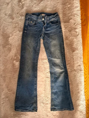 Blå bootcut jeans med slitningar - Säljer ett par blå bootcut jeans med snygga slitningar och klassisk femficksdesign. Jeansen har låg midja, bälteshällor och ljusare tvättade partier framtill. Perfekta för dig som gillar en avslappnad men trendig look.