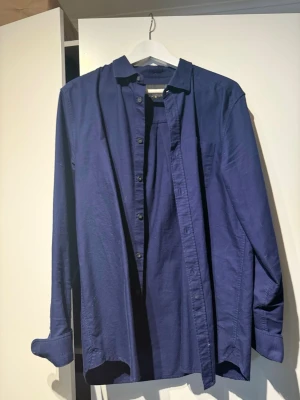 Marinblå skjorta från Lyle & Scott - Snygg marinblå skjorta från Lyle & Scott i klassisk modell med knappar framtill och bröstficka. Skjortan har lång ärm och är tillverkad i ett mjukt bomullstyg som känns skönt mot huden. Perfekt att styla lager på lager eller bära som den är.