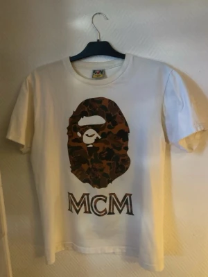 Vit MCM x Bape camo t-shirt - Vit t-shirt med stort camo-tryck av Bape-apan och MCM-logga på bröstet. Klassisk passform och rund halsringning. Perfekt för dig som gillar streetwear och vill sticka ut med en unik design. Den har blivit jättelite missfärgad vid krafen pga solkräm men det är inget man märker