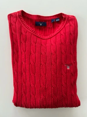 Röd kabelstickad tröja från Gant - Snygg röd kabelstickad tröja från Gant med rund halsringning och diskret logga broderad på bröstet. Tröjan har långa ärmar och ribbade muddar vid ärmslut och nederkant. Perfekt för dig som vill ha en klassisk och stilren look.