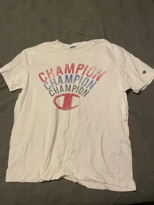 Vit Champion t-shirt  - Vit t-shirt från Champion med korta ärmar och stort tryck i rött och blått framtill där det står 'CHAMPION' flera gånger. Liten Champion-logga på ärmen. Klassisk passform och rund halsringning. Perfekt för en avslappnad stil.