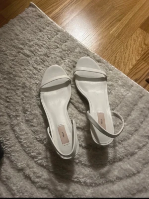 Vita klackskor  - Stilrena vita pumps från Nelly med öppen tå och smal rem över foten samt en justerbar ankelrem. Skorna har en klassisk, minimalistisk design och är tillverkade i ett slätt material som ger en elegant look.