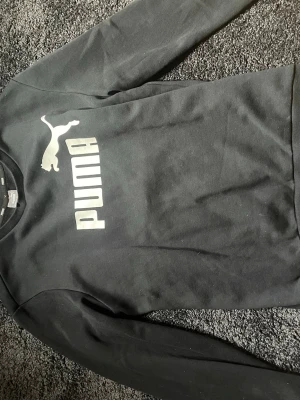 Svart sweatshirt från Puma - Svart sweatshirt från Puma med stort vitt logotryck på bröstet. Klassisk rund halsringning och långa ärmar. Tillverkad i mjukt bomullsmaterial för en bekväm och avslappnad stil.