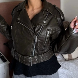 Svart biker skinnjacka med bälte - Snygg svart biker skinnjacka med bred krage, silvriga dragkedjor och detaljer. Jackan har ett matchande bälte i midjan och axelklaffar för extra attityd. Perfekt för dig som vill ha en edgy och trendig look.