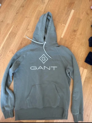 Grön hoodie från GANT med logga - Snygg grön hoodie från GANT med stor vit logga och emblem på bröstet. Klassisk känguruficka framtill och justerbar huva med dragsko. Tillverkad i mjuk bomullsblandning som känns skön mot huden. Perfekt för en avslappnad och trendig look.