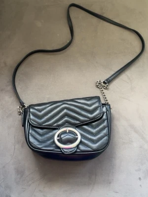 Svart quiltad axelväska med kedja - Snygg svart axelväska i quiltat konstläder med zigzag-mönster. Väskan har ett långt axelband med kedjedetalj och ett stort silverfärgat spänne framtill. Perfekt storlek för det viktigaste och ger en trendig touch till din outfit.