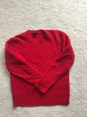Röd kabelstickad tröja Polo Ralph Lauren - Snygg röd kabelstickad tröja från Polo Ralph Lauren med rund halsringning och diskret broderad logga på bröstet. Tröjan har långa ärmar och ribbade muddar vid ärmslut och nederkant. Perfekt för dig som gillar klassisk stil.
