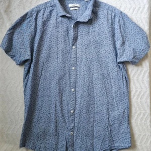 Blå kortärmad skjorta Jack & Jones - Snygg blå skjorta från Jack & Jones med diskret blommigt mönster. Skjortan har korta ärmar, knappar framtill och en klassisk krage. Slim fit-modell i mjuk bomull, perfekt för en avslappnad men stilren look.