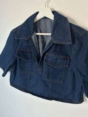 Kort mörkblå jeansjacka - Snygg kort jeansjacka i mörkblå denim med stora bröstfickor och bred krage. Jackan har korta ärmar och markerade sömmar för en cool look. Perfekt att slänga över en t-shirt för en avslappnad stil.