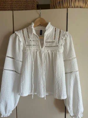 Vit boho-blus med spetsdetaljer. - Supersöt vit boho-blus från H&M med volangkrage, knytband och genombrutna spetsdetaljer på både ärmar och bröst. Blusen har en luftig passform och är tillverkad i ett strukturerat, lätt tyg som känns skönt mot huden. Jätte fin somrig bluss. Man kan antingen ha den öppen till halsen eller ha den knyten i en rosett.💗