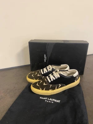 Saint Laurent svarta låga sneakers - Snygga låga sneakers från Saint Laurent i svart canvas med vitt grafiskt mönster. Skorna har vita snören, vit kant runt öppningen och en gulaktig gummisula. Perfekta för dig som vill ha något unikt och trendigt från ett lyxigt märke.39.5 men passar 40.5! 