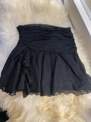 Svart kortkjol med volangdetalj - Säljer en svart kortkjol med volangdetalj på sidan och lätt rynkad midja. Kjolen har en flowy passform och är gjord i ett tunt, mjukt material som ger en snygg siluett. Perfekt för dig som gillar en stilren men ändå lekfull look.